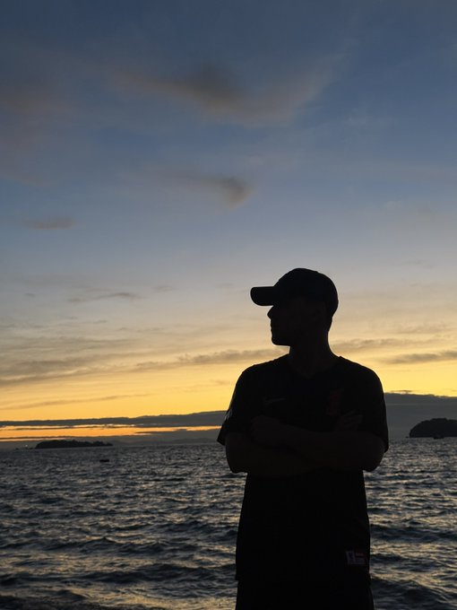 A silhouetted figure wearing a cap and jersey stands with crossed arms against a sunset sky over the ocean.