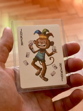 A hand holds a Joker playing card from the Bored Ape YC (BAYC) TCG, featuring a cartoonish ape character with a jester hat tossing cards, with "JOKER" printed vertically and "BAYC" along the right edge, showcasing the collectible gaming cards provided to holders.