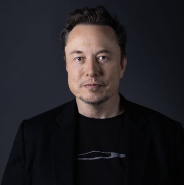 A man with short brown hair and a goatee, wearing a black blazer over a black t-shirt featuring a gray Tesla Cybertruck silhouette graphic, looks directly at the camera against a dark background. The image visually supports the post by featuring the Tesla logo, tying into the comparison between Tesla stock ($TSLA) and Amazon.