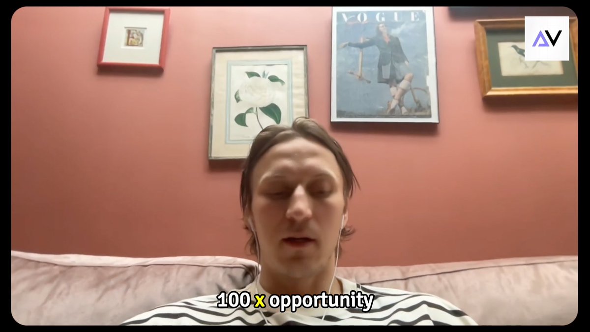 A video screenshot of a young man with long hair speaking on a couch, with captions reading "100 x opportunity" and an "AV" logo in the upper right corner, with framed Vogue and other artwork on a red wall behind him.