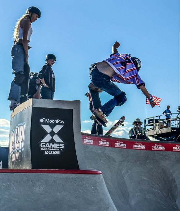A skateboarder in a red, white, and blue striped shirt performs a trick at the lip of a ramp during the X Games Sacramento 2026, with MoonPay branding prominently displayed on the obstacle and event signage. A helmeted skater and spectators, including an American flag, watch from the deck against a clear blue sky.