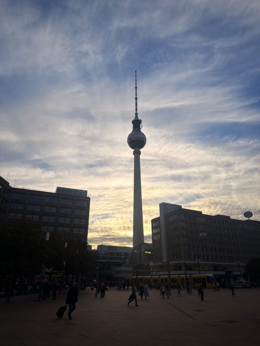 A view of the Berlin TV Tower (Fernsehturm) at dusk with people walking in the plaza below and a tram passing by.