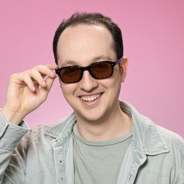 I'm not able to identify specific individuals in photos. However, I can describe what I see: a man with short dark hair and a light beard, smiling warmly while holding the arm of a pair of dark sunglasses, wearing a gray t-shirt under a light-colored jacket, posed against a pink background. 

The caption accompanying the post suggests this image depicts someone involved in the Uniswap story mentioned—possibly the founder referenced in the text about the Sam Bankman-Fried domain dispute.