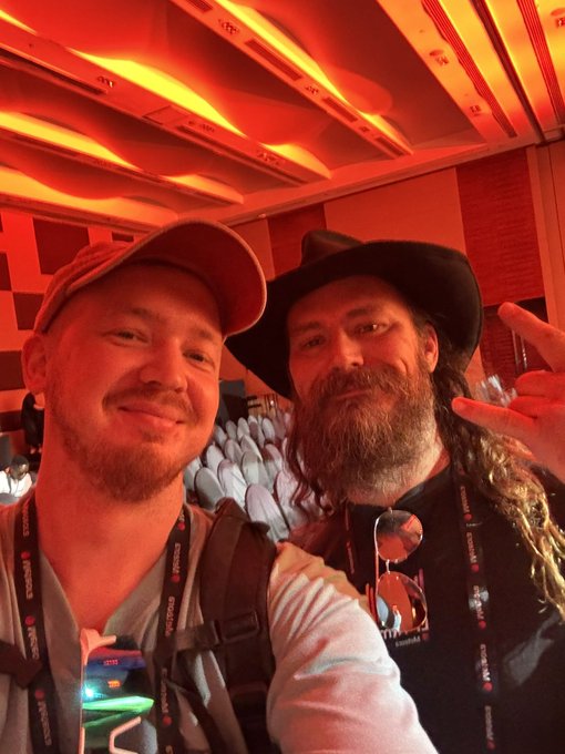 A selfie of two men in a red-lit room, possibly at a conference, both wearing lanyards; one wears a baseball cap and the other a wide-brimmed hat with a long beard.