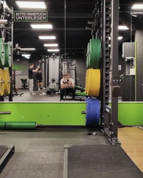 The image shows a gym with various exercise equipment, including green and yellow weight racks. There are people working out at the gym, some sitting on chairs while others lift weights or use other machines. The room has a black floor that contrasts with the colorful equipment. A person is seen using their cell phone in the middle of the scene, possibly taking a break from their workout to check messages or make plans.