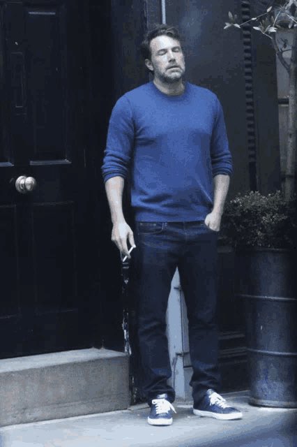The image features a man standing outside, dressed in blue jeans and a long-sleeved shirt. He has his hands in his pockets and appears to be smoking a cigarette. The background includes a door and a potted plant, adding context to the scene. There is also an umbrella visible in the image, which could potentially indicate that it's raining or about to rain.