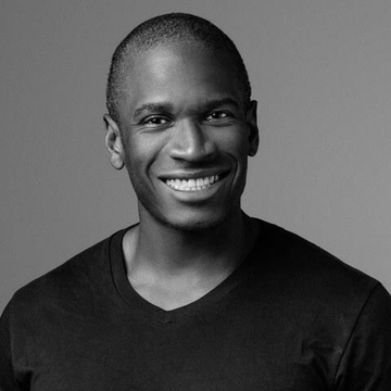 A black-and-white headshot of a smiling, bald man wearing a black v-neck t-shirt against a plain background.