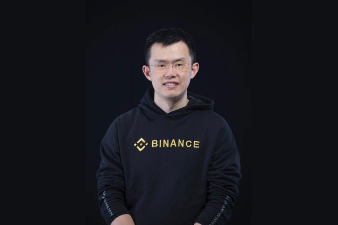 Professional portrait of CZ (Changpeng Zhao), former CEO of Binance, wearing glasses and a black Binance-branded hoodie against a dark background.