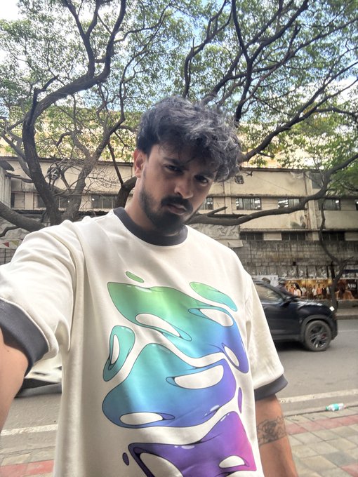 A selfie of a man with dark hair and beard wearing a white sweatshirt featuring a large abstract teal, green, and purple graphic, taken outdoors on a street with trees and a black SUV in the background.