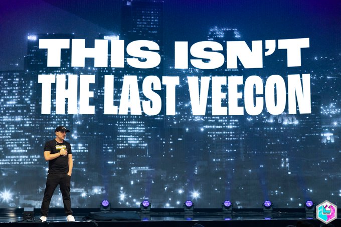 A man in black attire and a cap stands on a stage in front of a large screen displaying 'THIS ISN'T THE LAST VEECON' against a nighttime city skyline backdrop.
