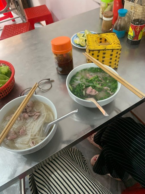 Two bowls of Vietnamese noodle soup sit on a metal table with chopsticks, condiments, and a basket of lime wedges.