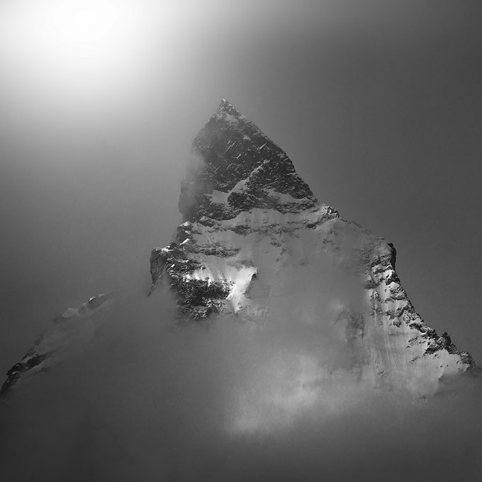 A black-and-white photograph of a snow-capped mountain peak, resembling the Matterhorn, emerging through thick clouds with sunlight glowing in the upper left corner.