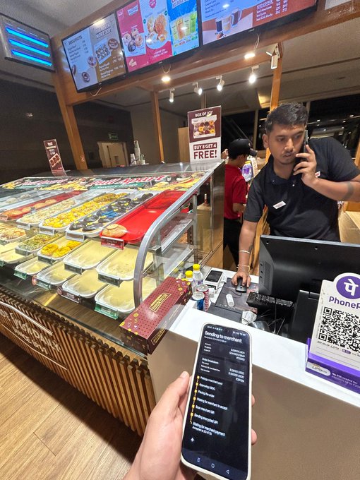 A person holds a smartphone showing a menu at a dessert/ice cream shop counter where a staff member is on the phone, with a QR code sign for PhonePe payment visible.