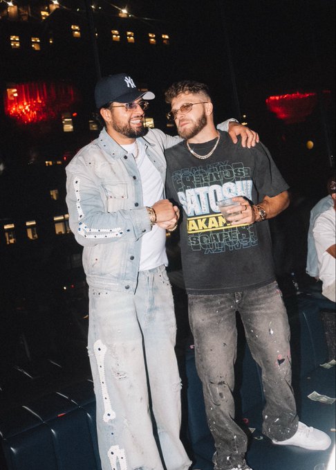 Two men posing together at a nighttime event, one wearing a Yankees cap and denim outfit, the other in a vintage-style graphic t-shirt and distressed jeans.