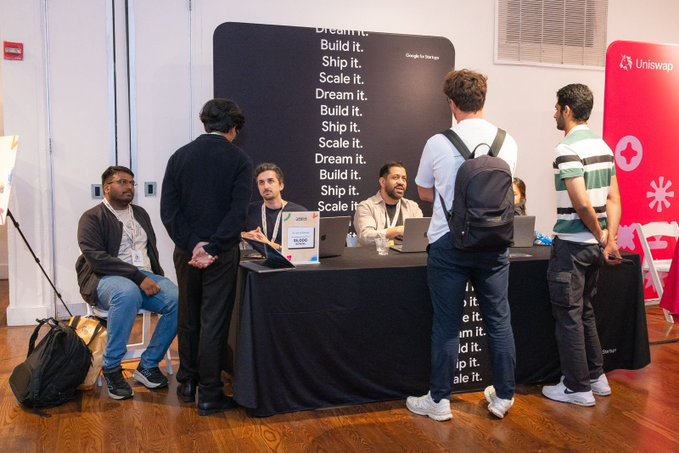 The image shows a group of people gathered around a table at an event, with some standing behind a desk while others are sitting down. The main focus of the scene is a large blackboard displaying inspiring words such as "dream big", "scale it", and "build it". A man in a backpack stands next to the table, possibly waiting for his turn or discussing ideas with someone else at the event. There are also several people standing around the room, engaged in conversations or observing the ongoing activities.