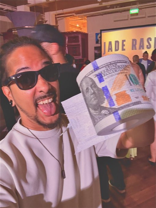 A man wearing sunglasses sticks out his tongue while holding up a roll of toilet paper printed with a $100 bill design, taken at an indoor event with a "JADE RAS..." sign partially visible in the background.