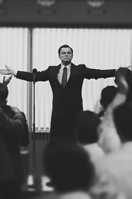 A black-and-white photograph of Leonardo DiCaprio in a pinstripe suit standing with arms outstretched in front of an audience, appearing to be a scene from The Wolf of Wall Street.