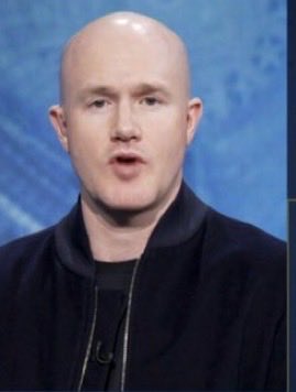 A bald man in a dark zip-up jacket speaks against a blue studio background.