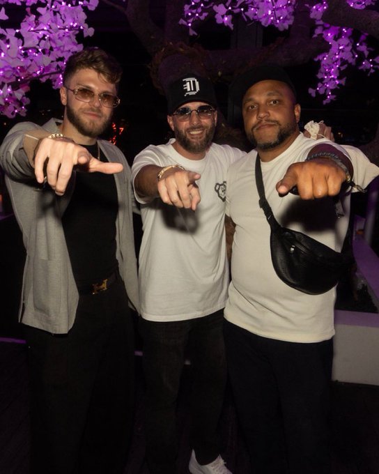 Three men pose for a photo at a venue with purple-lit cherry blossoms, smiling and pointing at the camera while wearing casual streetwear and caps.