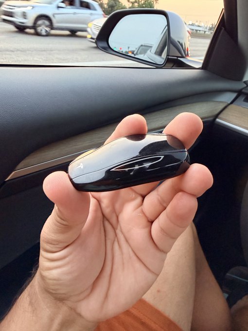 A hand holds a small, black, oval-shaped device resembling a Tesla key fob while sitting in a car with other vehicles visible through the window.