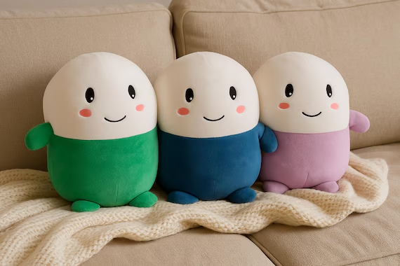 Three adorable egg-shaped plush mascots with cheerful smiles—one in green, blue, and pink—sit snugly together on a knit blanket, perfectly embodying the "Cupsey" character referenced in the post as a collectible mascot tied to the meme coin.