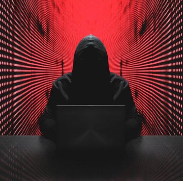 A shadowy hooded figure sits at a laptop in a dark environment, illuminated by radiating red and black digital lines that create a dramatic, ominous hacker-themed backdrop.
