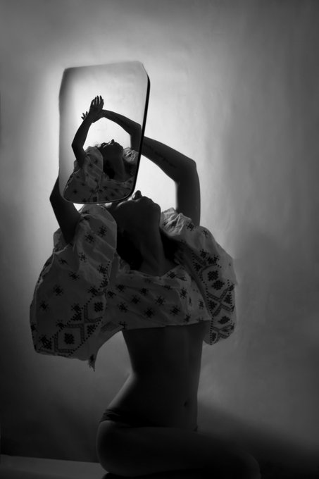 A black and white artistic photograph of a person holding a mirror up to their face, creating a surreal self-reflection effect.