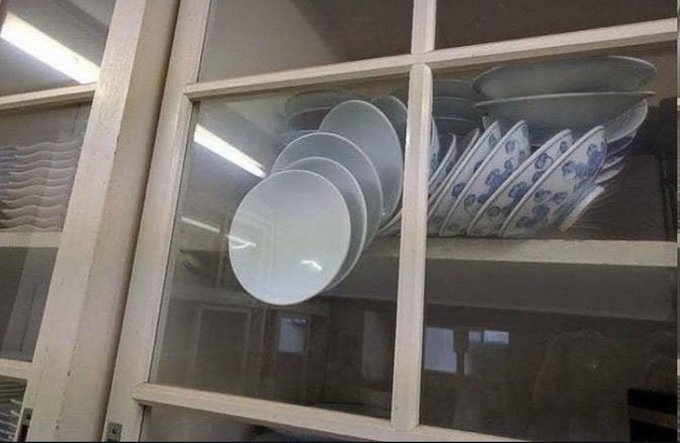 A cabinet displays neatly stacked white bowls and blue-and-white patterned plates on its shelves, creating a meme that humorously suggests Bitcoin is "stacked" and waiting, just like the orderly dishware lined up behind the glass.
