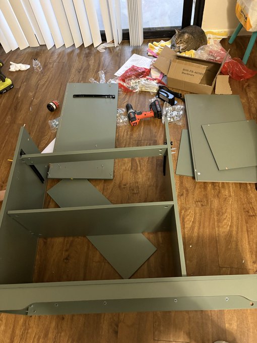 An indoor scene showing furniture assembly in progress, with green painted wooden panels and shelf pieces laid out on a wood floor, alongside tools including an orange drill, screws, hardware packaging, a cardboard box labeled 'B06-F8-41MM,' and a tabby cat observing from the background.