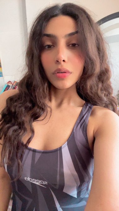 A selfie of a young woman with long wavy brown hair wearing a gray Adidas tank top.