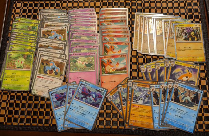 A large collection of Pokémon holographic cards is spread out on a woven mat, featuring a variety of characters including Charmander, Costume Pikachu, Komala, and Kricketot with visible HP values ranging from 50 to 180. The arrangement showcases numerous rare and shiny holographic cards, aligning with the "Cosmos holos" theme by highlighting a cosmic array of prized Pokémon collectibles.