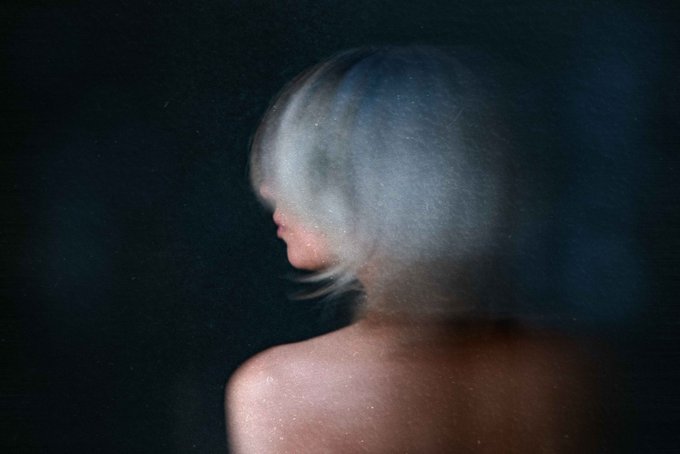 A figure with platinum blonde hair stands with their face turned away, partially obscured by a soft, misty blur that dissolves their features into the dark background. The ethereal, hazy quality mirrors the series' theme of "Beneath the Noise," visualizing the shedding of fear and expectation to reveal the quiet, authentic self beneath.