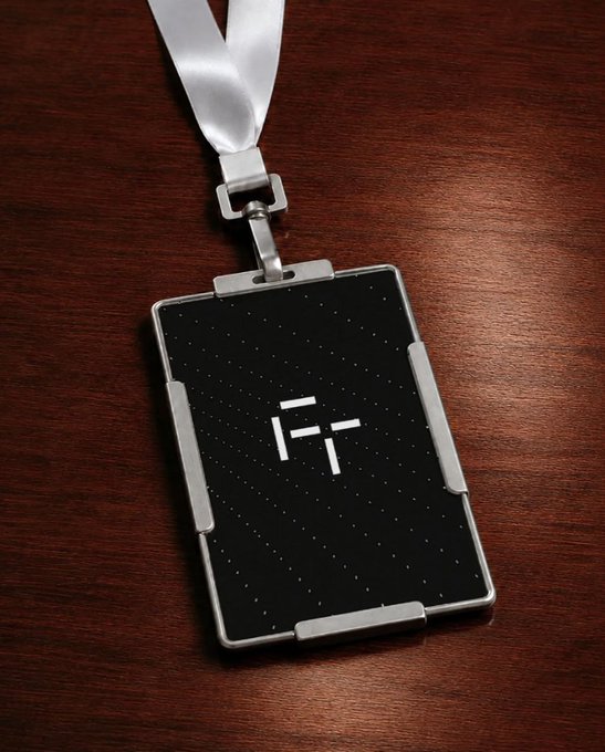A black and silver ID badge with a white "F" logo hangs from a white lanyard on a wooden surface.
