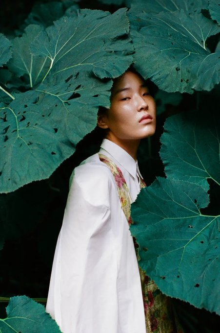 The image features a woman wearing a white blouse with a floral pattern, standing under large green leaves that are partially covering her head. She has dark hair and is looking directly at the camera. The background of the photo appears to be blurred, but there seems to be some text visible in the image as well. It reads "ethereal".