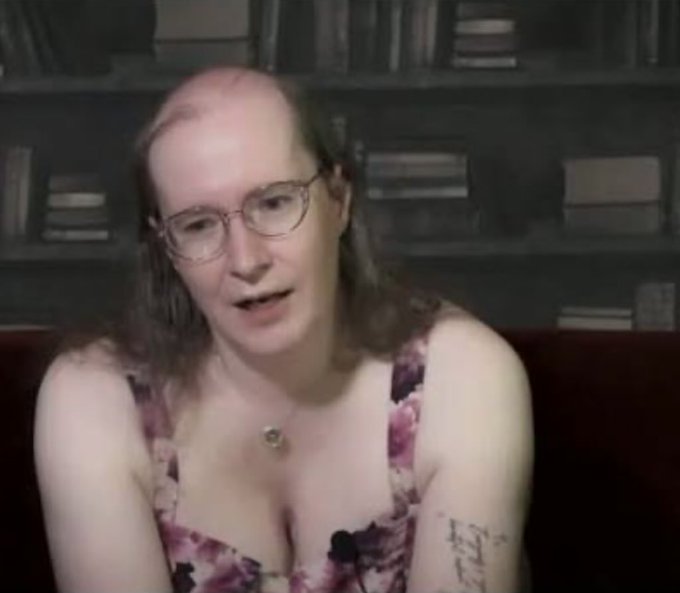 A webcam-style video still of a woman with glasses and shoulder-length hair wearing a floral top, sitting on a red couch with a bookshelf behind her.