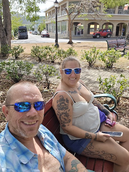 A selfie of a smiling couple wearing sunglasses sitting on a park bench, with the woman displaying arm and leg tattoos and holding a phone, while buildings, a bus, and a Jeep are visible in the background.