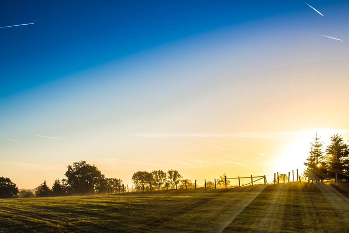 A serene landscape showing a sunset over a grassy hill with silhouetted trees and a wooden fence, under a blue sky streaked with light trails.