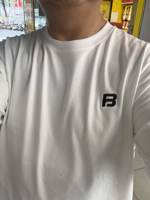A selfie showing the upper torso of a person wearing a white sweatshirt with a dark blue 'B' logo, taken in front of a storefront.