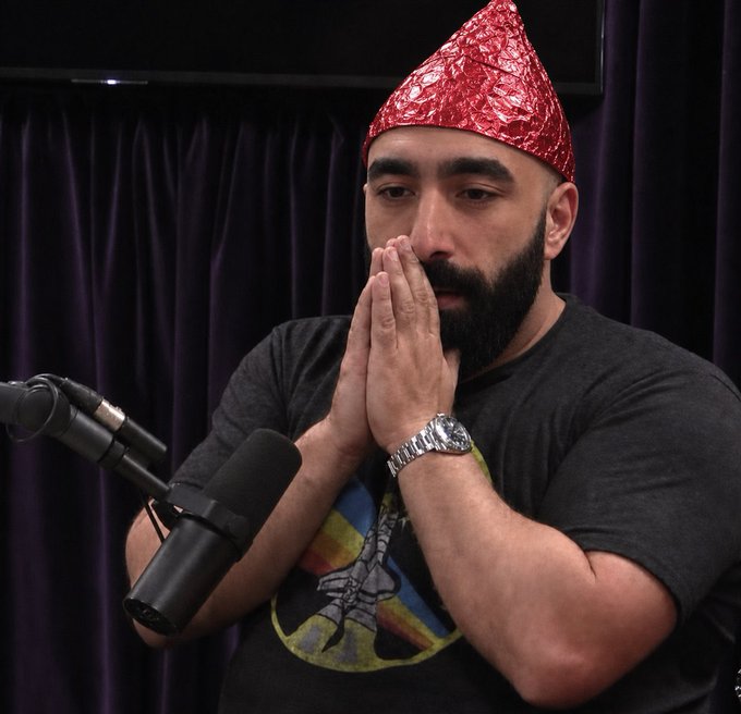 A bearded man wearing a red foil hat sits before a microphone with his hands clasped in a prayer-like gesture, wearing a watch and a graphic t-shirt. The image appears to be from a podcast setting, with the post referencing Anatoly Yakovenko (known as "Toly"), co-founder of Solana (SOL), suggesting the man is humorously praying for Turkey to beat the United States in a sports match so he will swap his SOL holdings into AVAX.