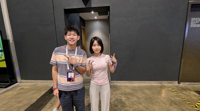 A young man and woman pose making hand gestures in a modern building interior, with a visible sign mentioning 'Company Exit, Premises CCTV in Operation'.