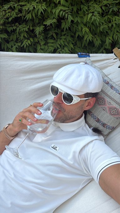 A man in a white cap, white sunglasses, and a white Lacoste polo shirt reclines on an outdoor daybed, drinking from a wine glass filled with water, wearing a gold bracelet and a green ring.