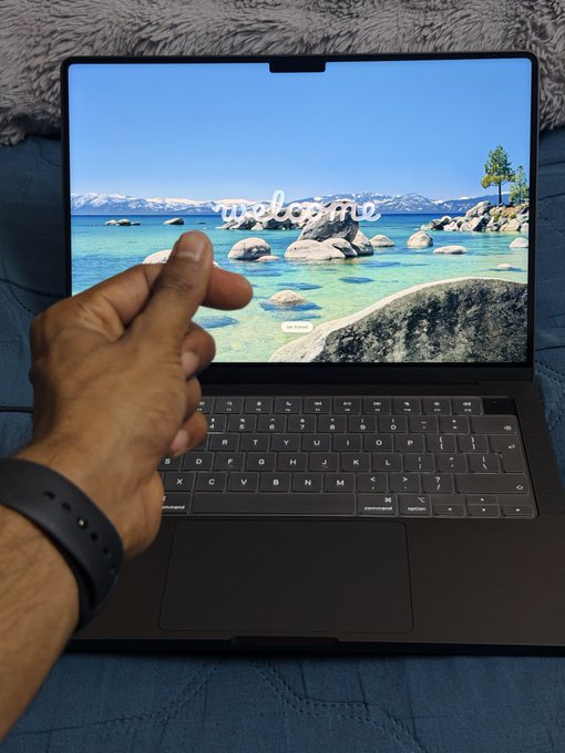 A hand makes a finger heart gesture over a laptop keyboard, with a scenic wallpaper displaying "Welcome" on the screen.