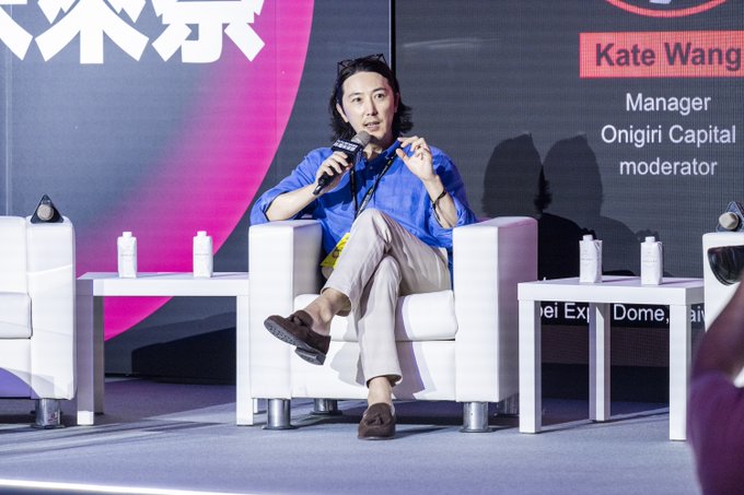 A photograph of Kate Wang, Manager and moderator from Onigiri Capital, speaking into a microphone while seated in a white armchair on a stage at a panel event.