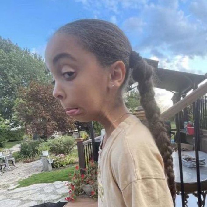 A side profile of a young woman with long braided hair in a ponytail, standing outdoors in a residential area.