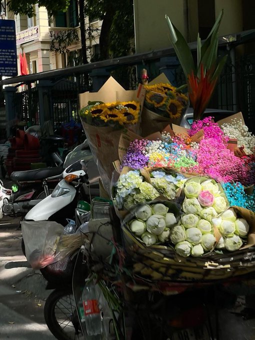 The image features a street scene with various items for sale. There are two motorcycles parked next to each other, one of which has flowers displayed on its back. The other motorcycle is empty. In front of these motorcycles, there's an assortment of colorful flowers in baskets and bags, including sunflowers, carnations, and roses. A bicycle can also be seen nearby.

The post accompanying the image provides information about a flower shop located at GM cả nhà Một ngày mình Cứ đều đặn làm những gì mình, and the address of the flower shop.