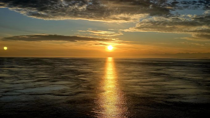 A golden sunset reflects across the calm waters off the Alaskan coast, with no grass or land visible—just endless ocean and a fiery horizon mirroring the user's "Alaska views good tho" sentiment.