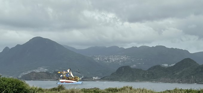A scenic landscape view showing a harbor with a boat in the foreground and mountainous terrain under a cloudy gray sky.