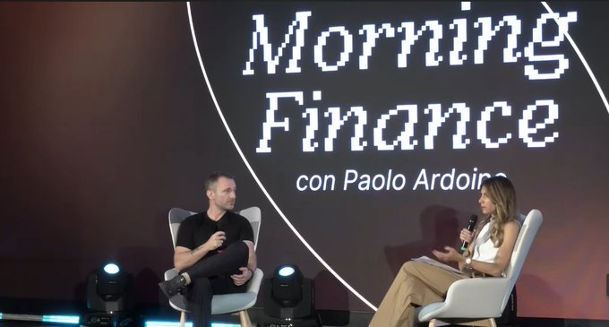 A man and woman sit in chairs on stage with microphones during a "Morning Finance con Paolo Ardoino" panel discussion. The setting aligns with the post about an interview with Silvia Berzoni at "The Last Question" event discussing Tether's mission and its critique of the traditional financial system.