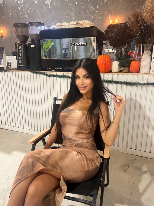 A woman in a satin brown dress sits in a director's chair in front of a counter with coffee equipment (Conti, Nitro), pumpkins, and dried flowers.