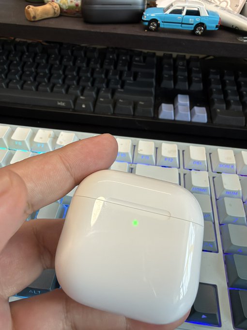 A hand holding white wireless earbuds case with a green indicator light, positioned in front of two keyboards.